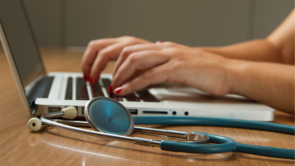 A women typing on a laptop keyboard, on a wooden table, with a blue stethoscope on the side.